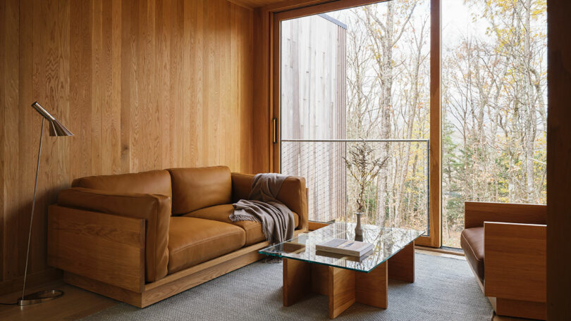 Modern living room with wood-paneled walls, a Design Within Reach brown leather sofa, glass coffee table, wooden armchair, floor lamp, and large window overlooking trees.