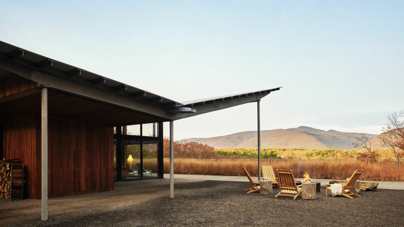 Modern wooden house with large windows and overhanging roof; Design Within Reach-inspired outdoor seating area with a fire pit; mountains and trees in the background.
