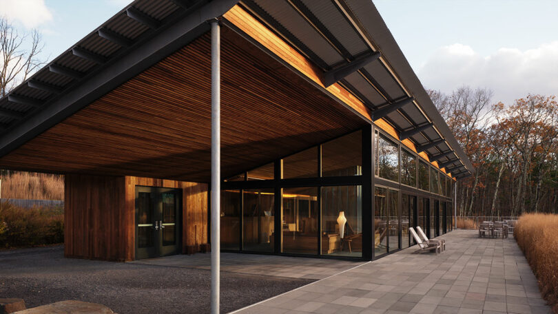 Modern house with a sloped metal roof, large glass walls, wooden accents, and a paved patio featuring Design Within Reach outdoor chairs, surrounded by dry grass and trees.