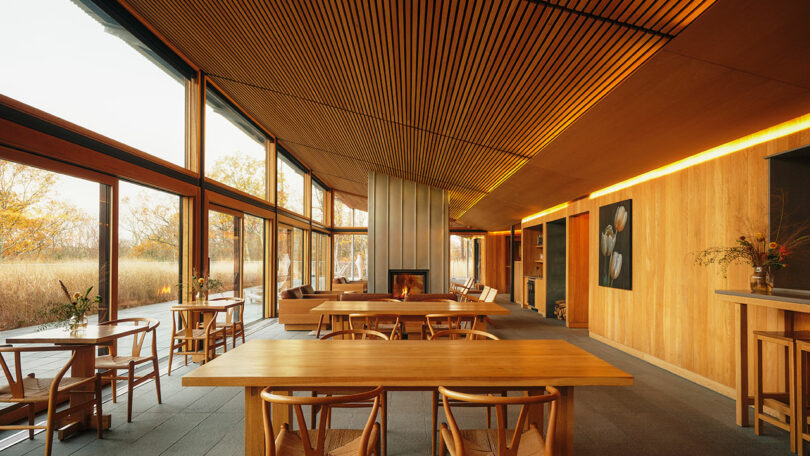 Modern restaurant interior featuring Design Within Reach wooden tables and chairs, large floor-to-ceiling windows, wood-paneled walls, and a central fireplace, with natural light streaming in.