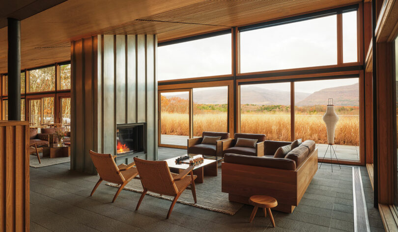 Modern living room with large windows, brown leather sofas from Design Within Reach, woven chairs, a central fireplace, and views of tall grass and distant mountains outside.
