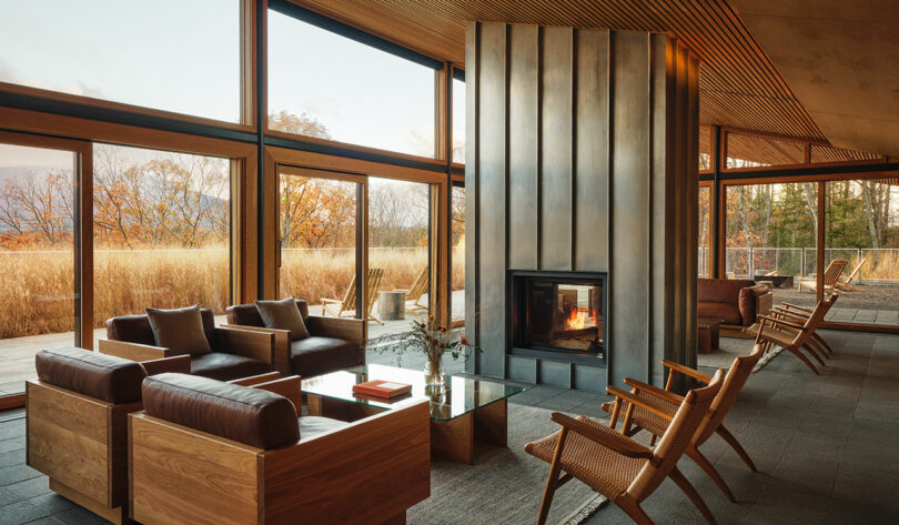 Modern living room with large windows, wooden furniture from Design Within Reach, a metal fireplace, and views of the autumn landscape outside.