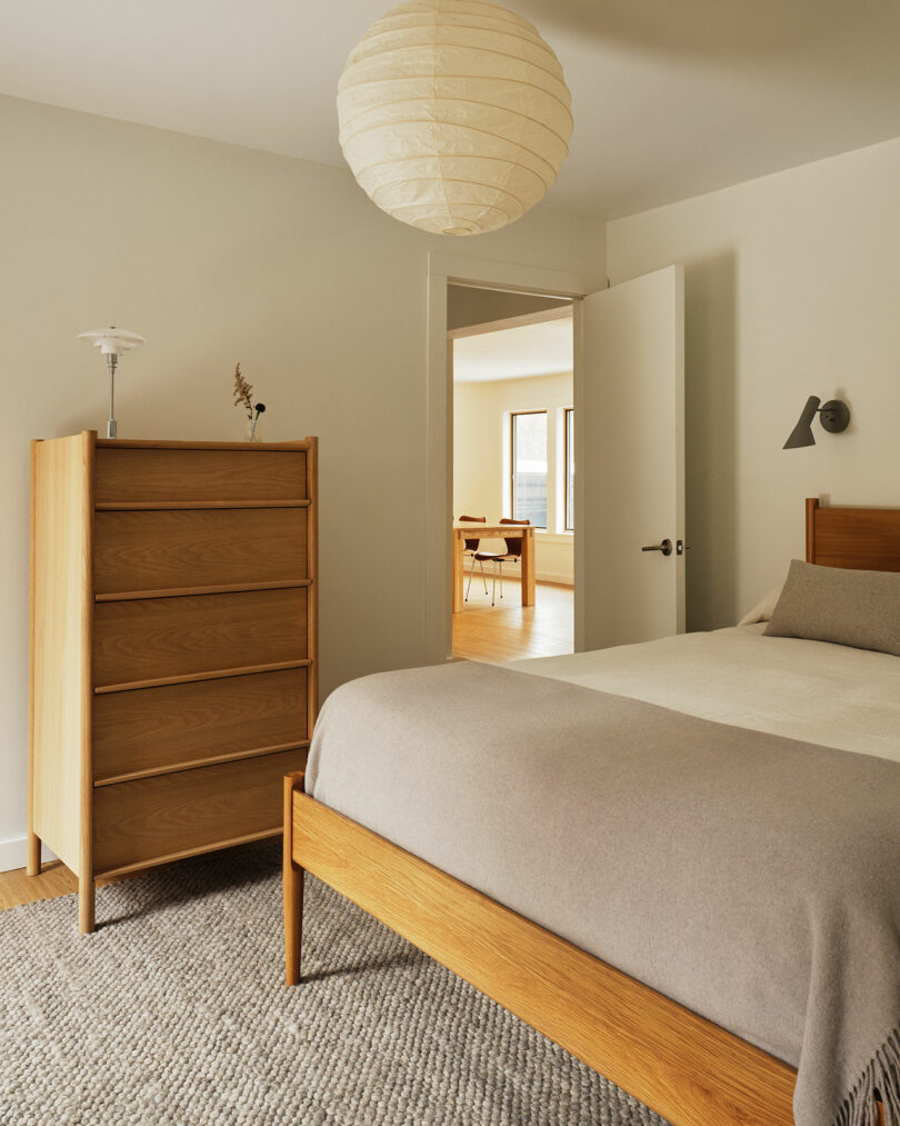 A minimal bedroom with a wooden bed and dresser, neutral bedding, a gray rug, and a round paper ceiling lamp—styled with Design Within Reach touches; an open door leads to a sunlit adjacent room.