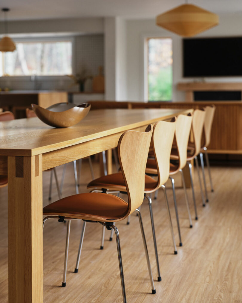 A row of wooden chairs with brown seats is lined up along a light wood dining table in a modern, well-lit dining area, echoing the sleek style of Design Within Reach.