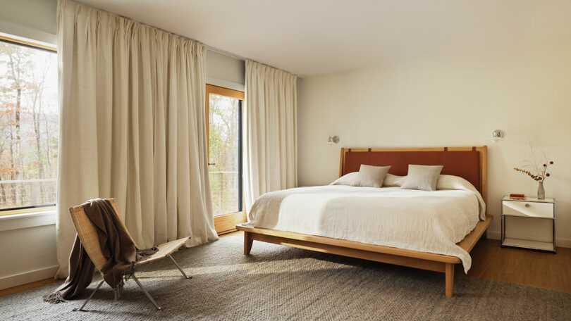 A minimalist bedroom with a large bed, neutral bedding, two pillows, a side table, a simple chair draped with a brown blanket, and light curtains framing the large windows—effortlessly stylish in the spirit of Design Within Reach.