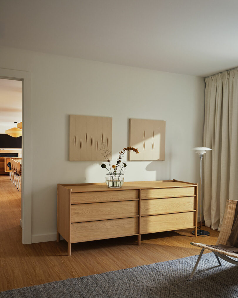 A minimalist room with two wooden dressers, two abstract beige artworks on the wall, a Design Within Reach glass vase with flowers, a floor lamp, and a woven chair on a gray rug.