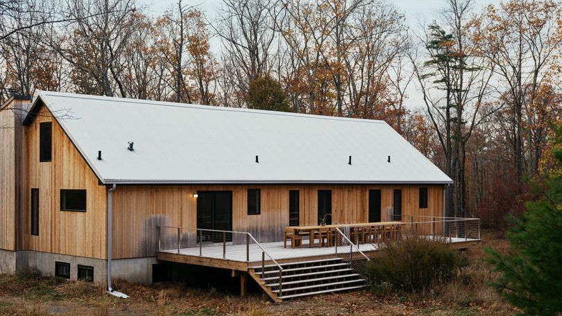 A modern wooden house with a metal roof, large deck, and outdoor seating inspired by Design Within Reach, surrounded by leafless trees in an autumn landscape.