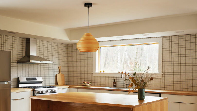 Modern kitchen with tiled walls, wooden island, gas stove with vent hood, large window, and minimalist decor including a vase with dried flowers—styled with touches inspired by Design Within Reach.