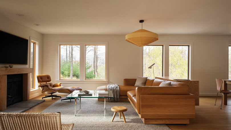 Modern living room with large windows, a tan leather sofa from Design Within Reach, glass coffee table, lounge chair with ottoman, and wooden pendant light. Neutral color palette enhanced by natural light.
