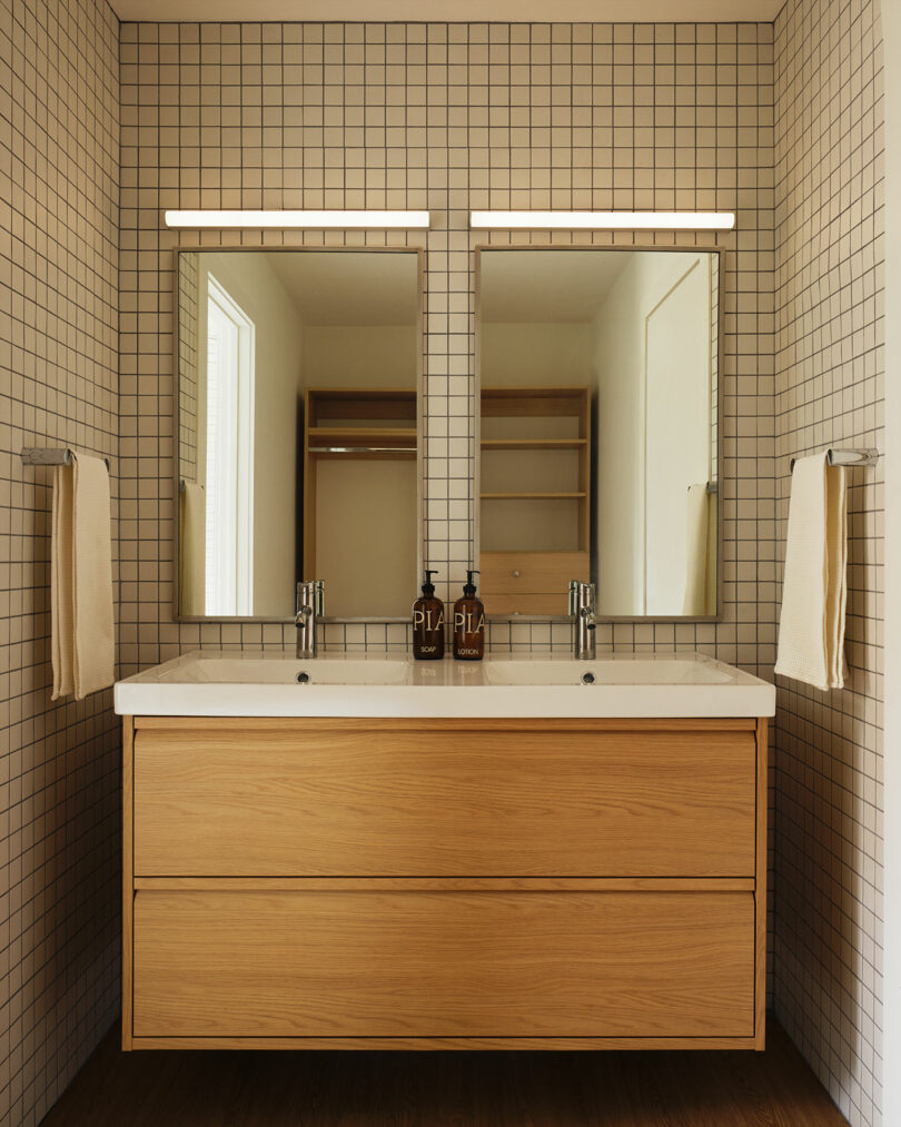 Modern bathroom with a double sink vanity, Design Within Reach mirrors, grid-tiled walls, wall-mounted lights, and beige towels on metal racks.