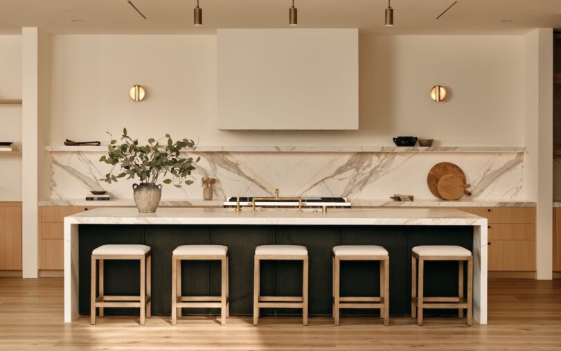Modern kitchen with a marble island, four stools, minimalist decor, light wood flooring, and built-in shelves. Neutral tones and clean lines create a sleek, contemporary look.