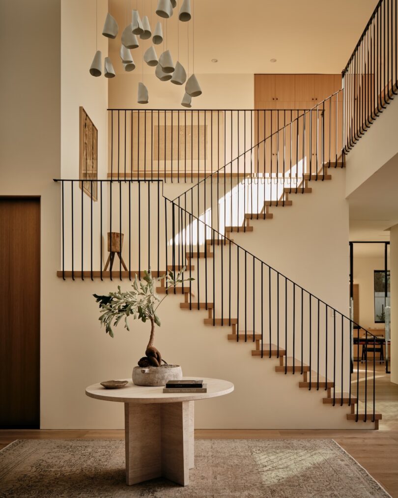 A modern foyer with a central round table holding a plant, neutral decor, a staircase with black railings, and pendant lights hanging from a double-height ceiling.