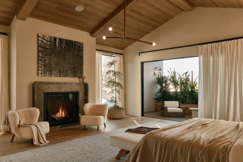 A cozy bedroom with a fireplace, two armchairs, a bed, and floor-to-ceiling curtains, opening to an outdoor patio with greenery and a bathtub.