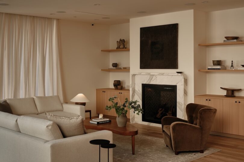 A modern living room with neutral tones, featuring a marble fireplace, built-in shelves with decor, a beige sofa, two brown armchairs, and a wooden coffee table.