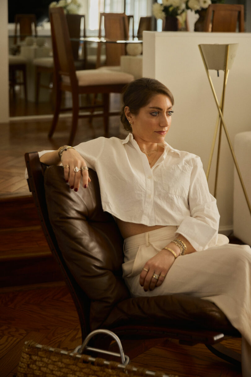Coco Greenblum sits in a brown leather chair, wearing a white blouse and light pants, in a modern, well-lit living room with wooden floors and dining furniture in the background.