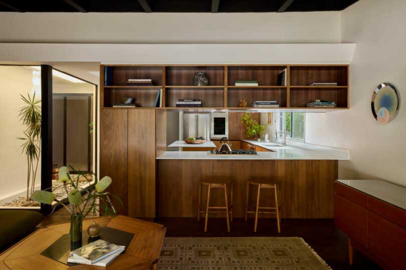 Modern kitchen with wood cabinetry, open shelves displaying books—including a Coco Greenblum cookbook—a white countertop, two wooden stools, and a wall-mounted round decorative plate.