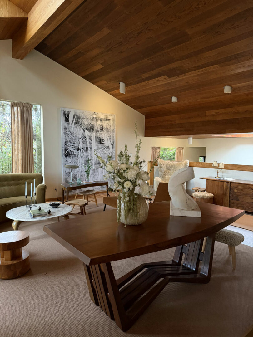 A mid-century living space with a wood-beamed ceiling, walnut dining table with floral arrangement, marble sculpture, abstract wall art by Coco Greenblum, and a green velvet sofa.