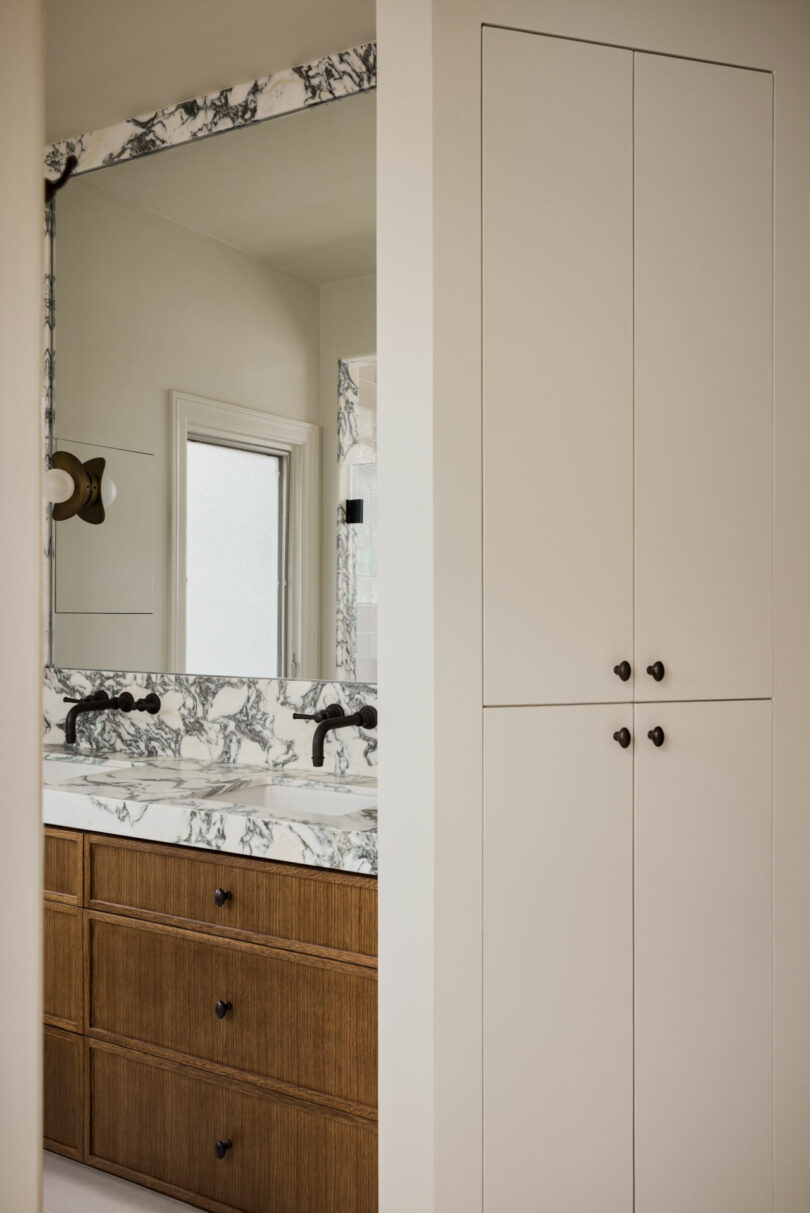 Modern bathroom with a large mirror, marble countertop and backsplash, wood vanity drawers, and a tall white storage cabinet with dark knobs—styled by Coco Greenblum.