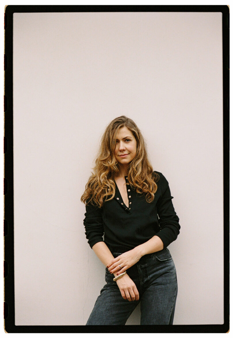 A woman with long, wavy hair stands against a plain light-colored wall, wearing a black buttoned shirt and dark jeans. She looks at the camera with a neutral expression.