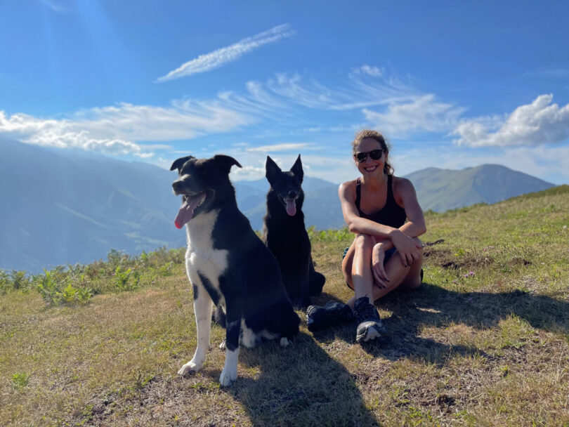 A woman in sunglasses sits on grass with two dogs on a sunny hillside, with mountains and a blue sky in the background.