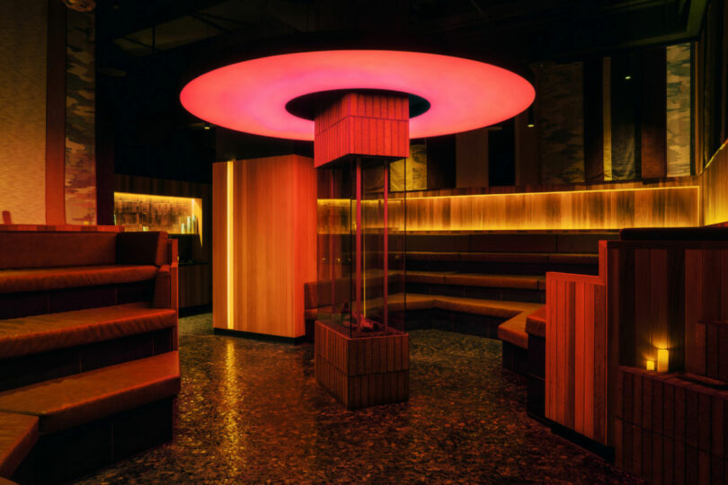 A modern lounge with tiered seating, wood-paneled walls, warm ambient lighting, and a central column topped by a glowing red circular light fixture.