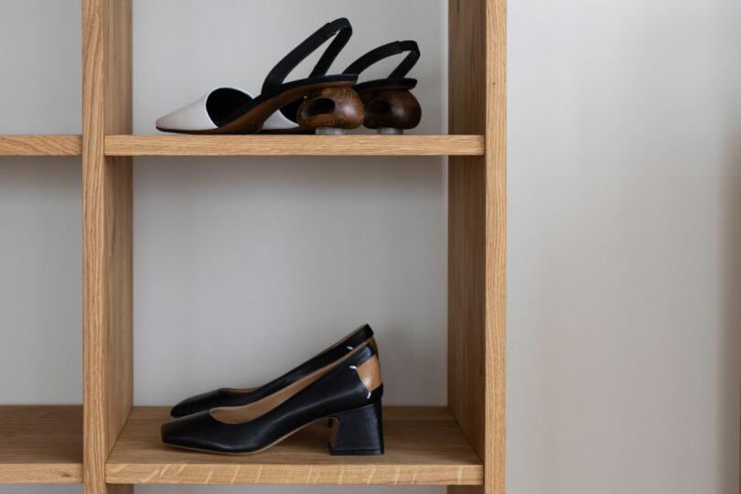 Two pairs of women's heeled shoes, one with wooden cutout heels and one with block heels, are displayed on adjacent shelves of a wooden shelving unit.
