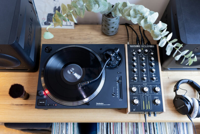 Overhead view of a turntable playing a vinyl record beside an audio mixer, headphones, and speakers on a wooden table. A potted plant and records are also visible.