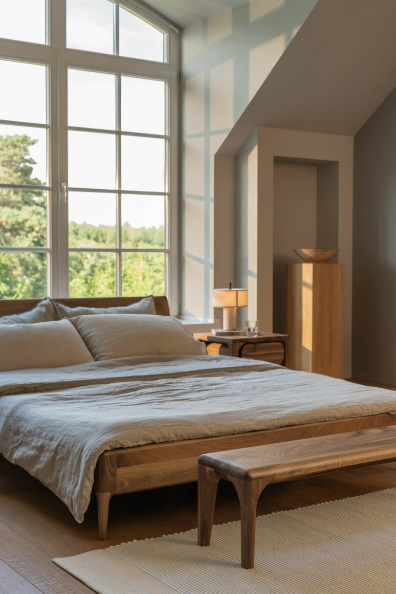 A minimalist bedroom with a large window, wooden bed, matching bench, neutral bedding, side table with lamp, and a decorative bowl on a pedestal. Natural light fills the room.