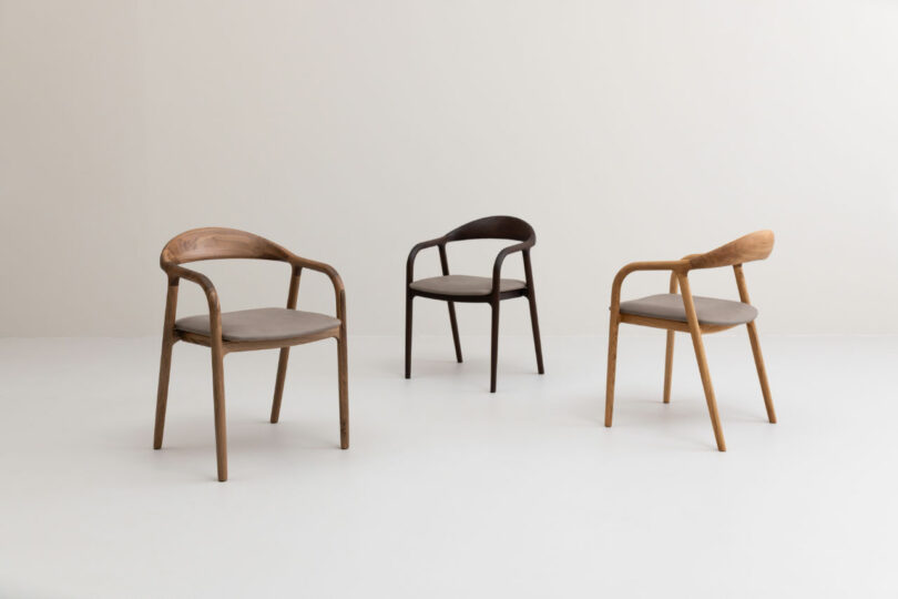 Three wooden chairs with curved backs and armrests, each in a different wood tone, are arranged on a white floor against a plain, light-colored background.
