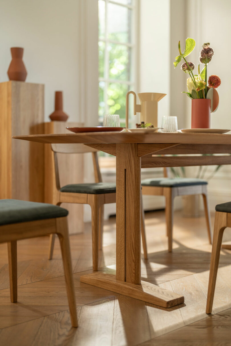 A wooden dining table with chairs, set with glassware, a ceramic pitcher, a plate, and a vase of flowers, in a bright room with wooden flooring and large windows.