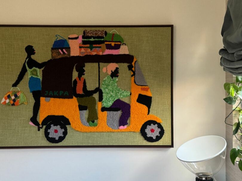 A textile artwork depicting three people riding in an orange auto-rickshaw with luggage on top, hung on a wall beside a white floor lamp and a plant.