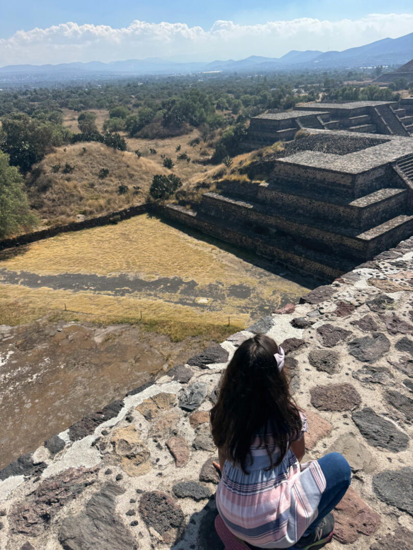 A person with long dark hair sits on a stone ledge overlooking ancient stone pyramids and a grassy area, with mountains visible in the distance.