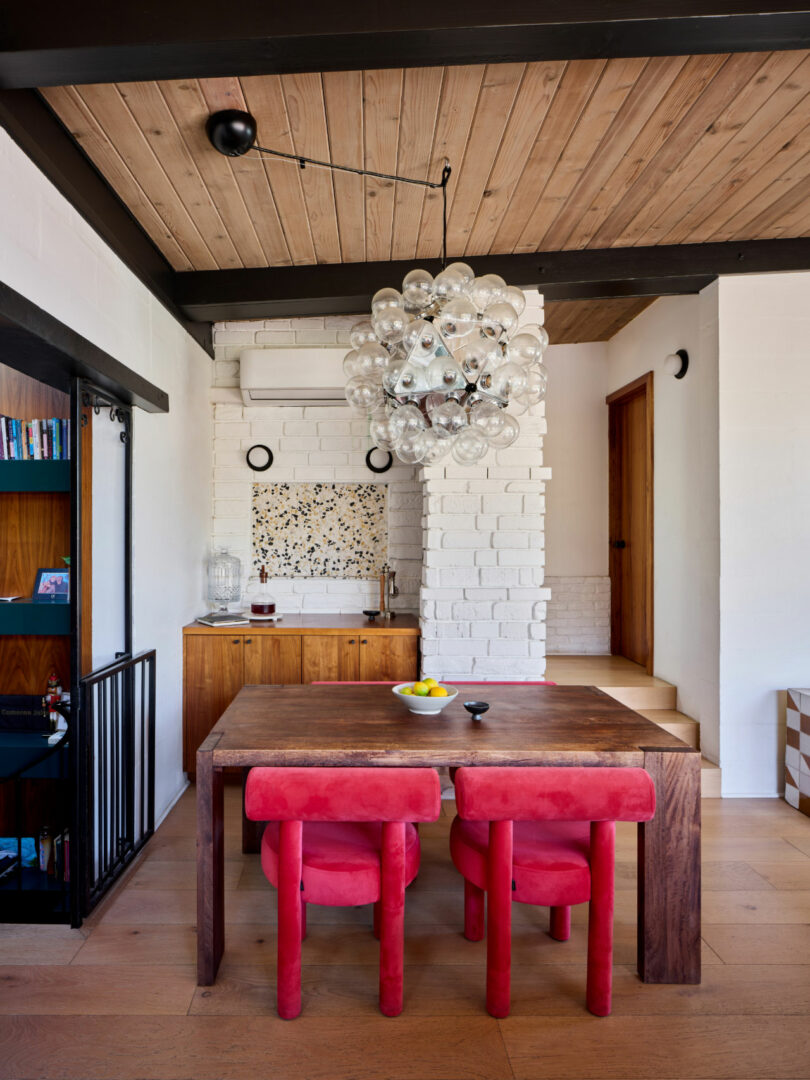 A modern dining area with a wooden table, four red chairs, a bubble chandelier, and a small kitchen area with wooden cabinets and white brick walls.