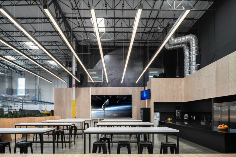 A modern industrial cafeteria with long tables, black stools, a kitchenette, wall art of a space scene, and exposed ceiling pipes.