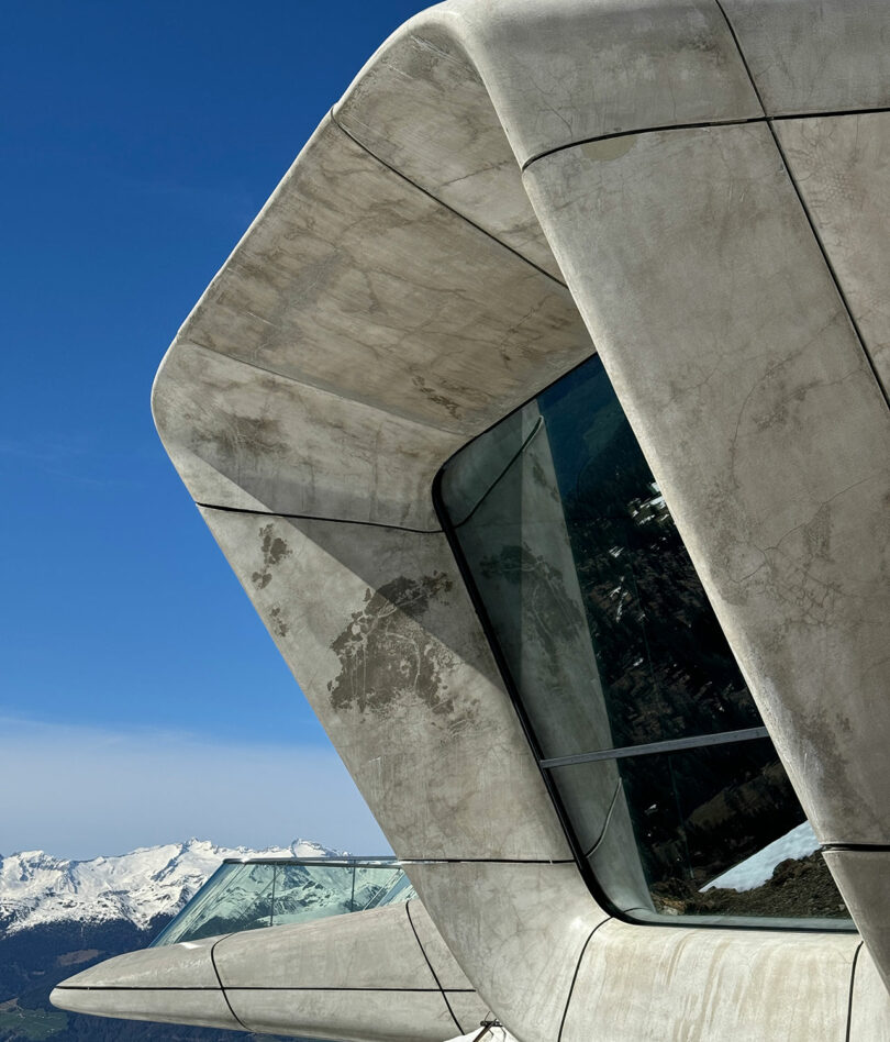 Close-up view of Kameh’s modern concrete building with angular architecture and large glass windows, set against a blue sky and snow-capped mountains in the background.