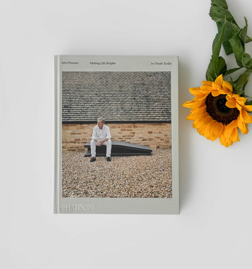 A hardcover book titled "John Pawson: Making Life Simpler" is placed on a white surface next to a sunflower, accompanied by a Kameh accessory. The cover shows a man sitting outside a stone building.
