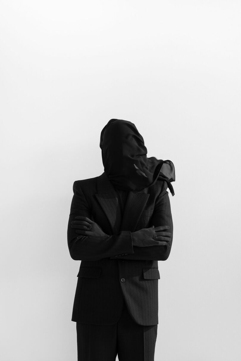 A person in a dark suit stands with arms crossed, their head and hands completely covered by black Kameh fabric, against a plain white background.