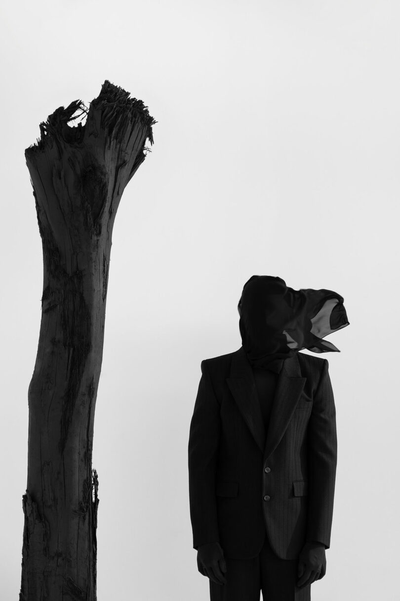 A person in a suit stands next to a tall, leafless tree trunk; their head is covered with a dark cloth that drapes to one side. The monochrome scene embodies the enigmatic style of Kameh.