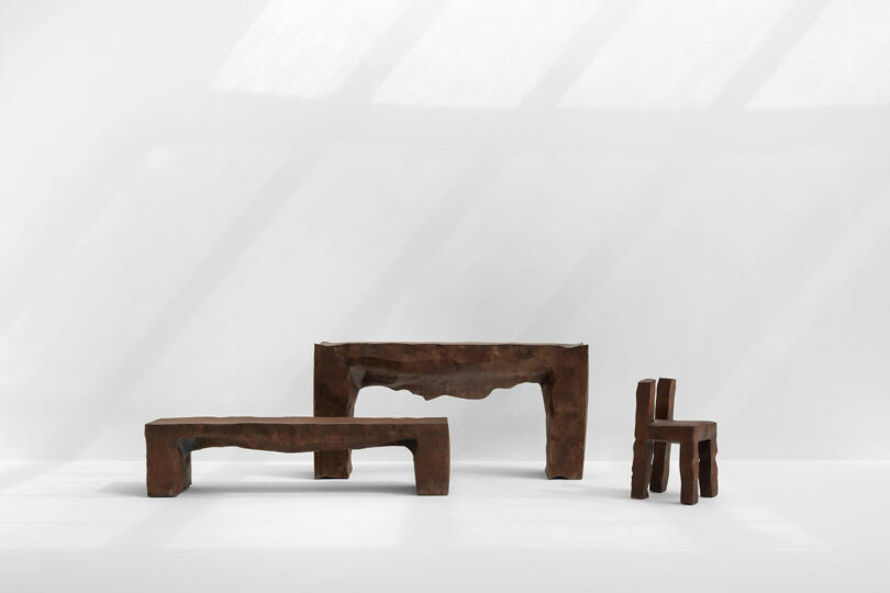 Three sculptural, rust-colored Kameh furniture pieces—a bench, a console table, and a chair—are displayed against a plain white wall under soft natural light.