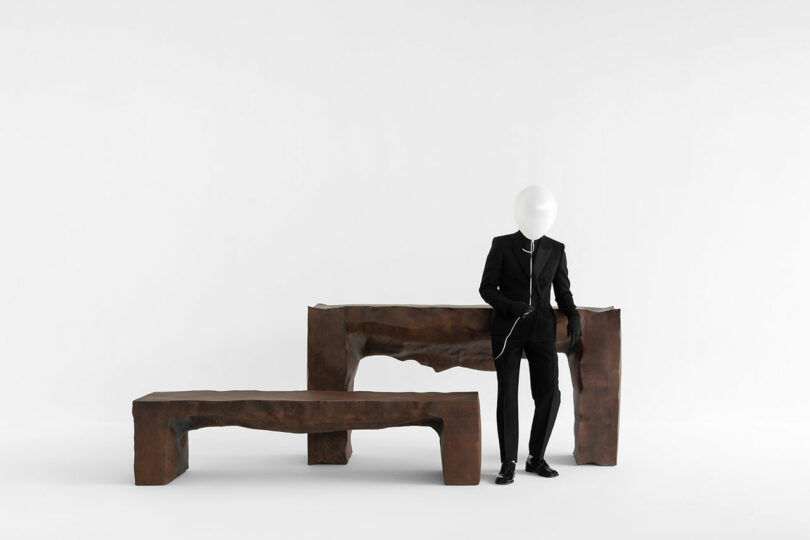 A person in a black suit with a white balloon covering their head stands beside two irregularly shaped brown Kameh benches against a plain white background.