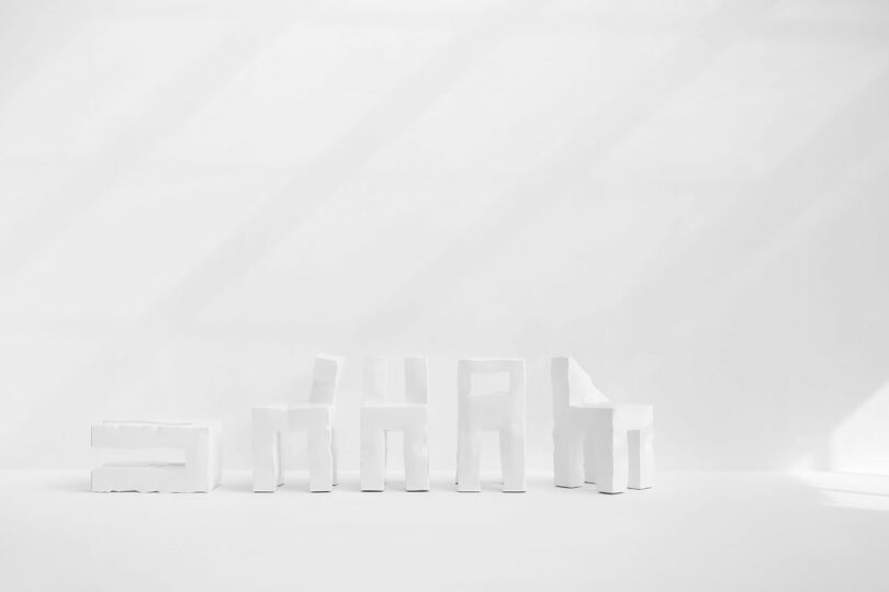 Six white Kameh geometric chairs are arranged in a row against a plain white background, with diagonal shadows cast on the wall.
