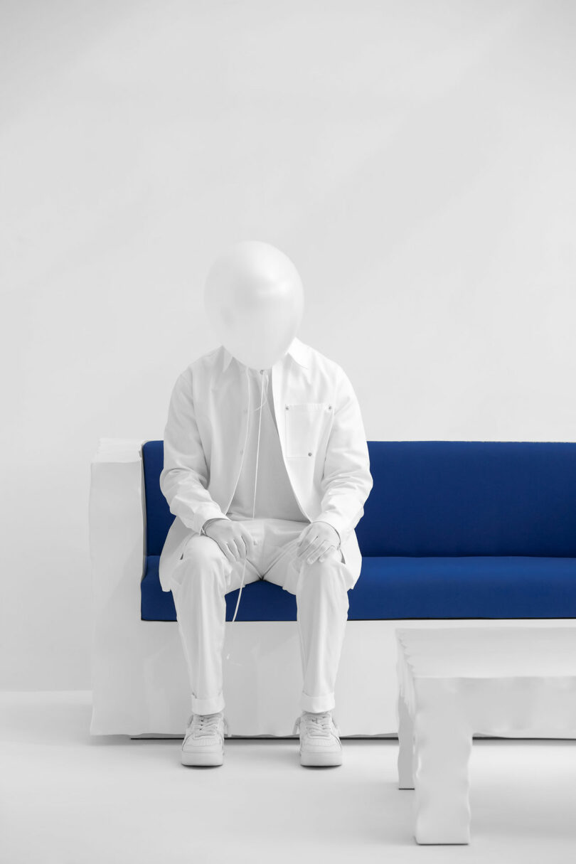 A person dressed in all white with a Kameh balloon instead of a head sits on a blue and white sofa next to a white table in a minimalist, white room.