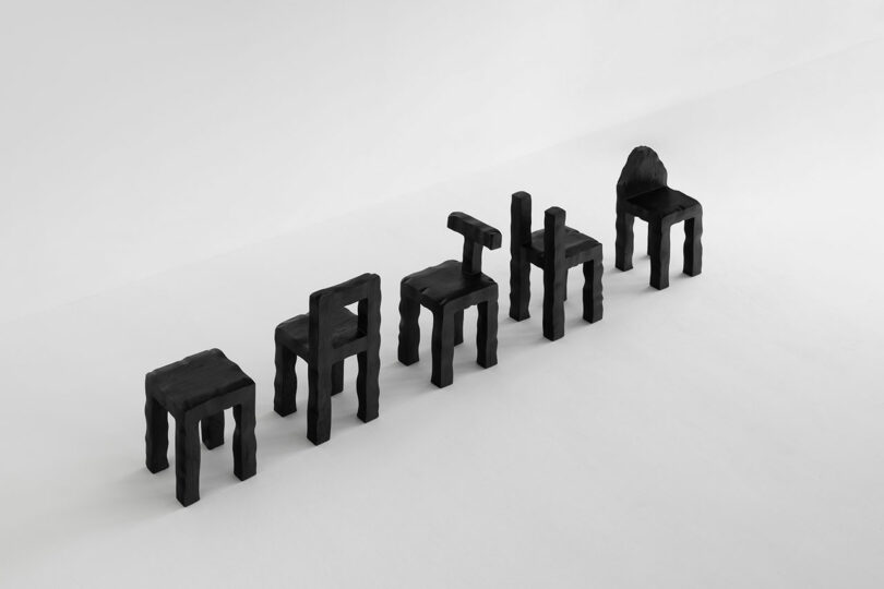 Six black Kameh chairs with blocky, irregular shapes are arranged in a row on a white floor, each designed to resemble a different letter.