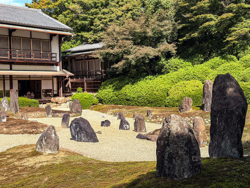A traditional Japanese rock garden with large and small stones arranged on gravel, next to a wooden building, surrounded by green shrubs and trees, inspired by artist Nimrod Weis.