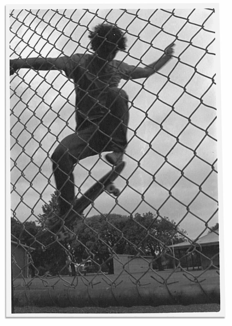 In this black and white photo, a person climbs a chain-link fence outdoors, with trees and buildings behind them—capturing a moment reminiscent of Nimrod Weis's evocative visual style.