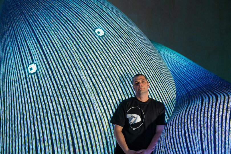 A man in a black shirt stands in front of a large, striped, abstract sculpture with two circular patterns resembling eyes.