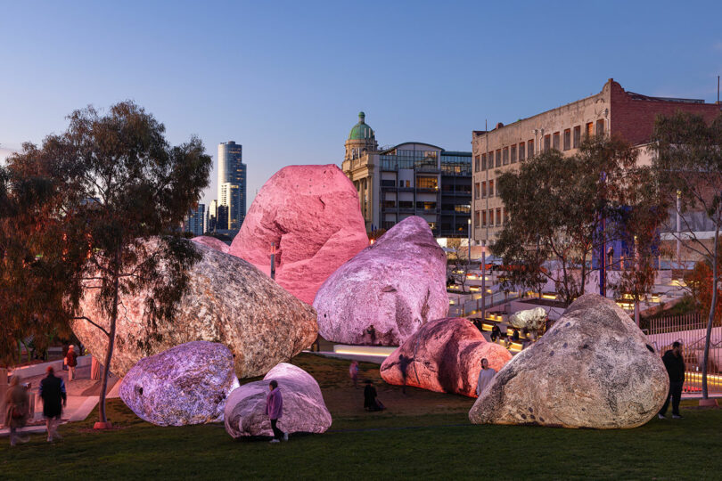 Large artificial boulders by Nimrod Weis, illuminated with pink and purple lights, stand in an urban park as people stroll nearby at dusk.