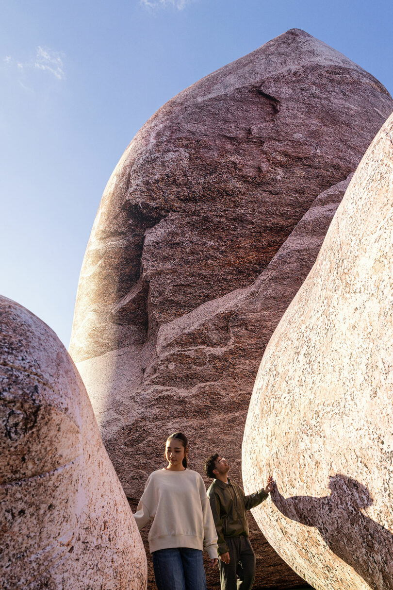Two people stand among large, smooth, reddish rock formations under a clear blue sky. One person is in the foreground, the other behind—both touching the rocks, exploring their textures like Nimrod Weis in an immersive natural landscape.