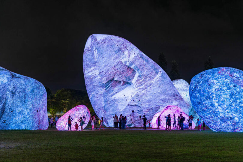 Large rock-like sculptures by Nimrod Weis illuminated in blue, purple, and pink lights at night, with groups of people standing and walking around them on grassy ground.