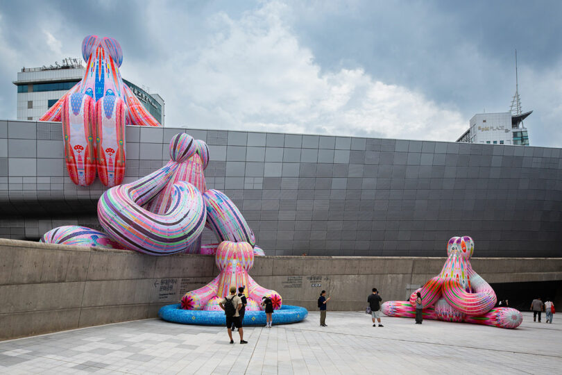 People photograph large, colorful inflatable sculptures by Nimrod Weis, resembling abstract creatures placed around a modern urban building under a cloudy sky.